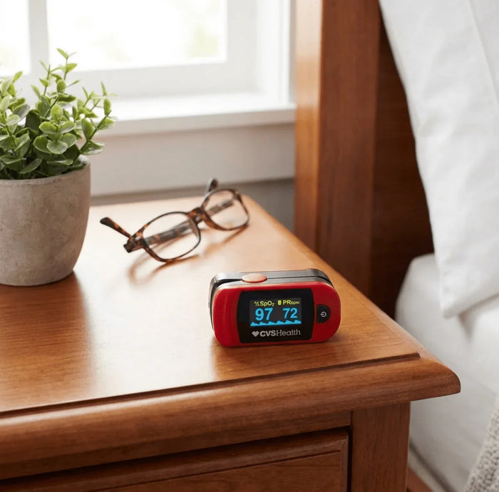Portable pulse oximeter displaying oxygen and heart rate on a bedside table with glasses.