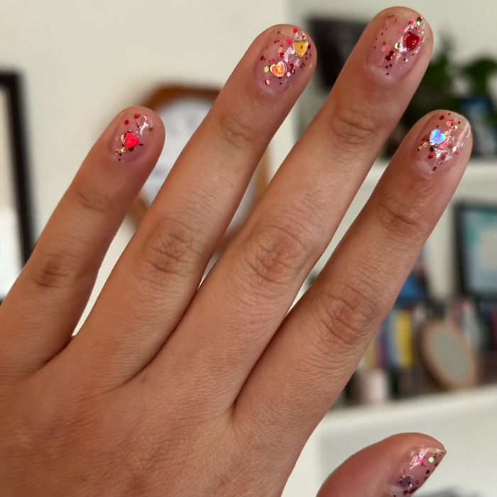 Hand with natural nails featuring HEARTED quick dry polish decorated with small hearts and sequin confetti manicure