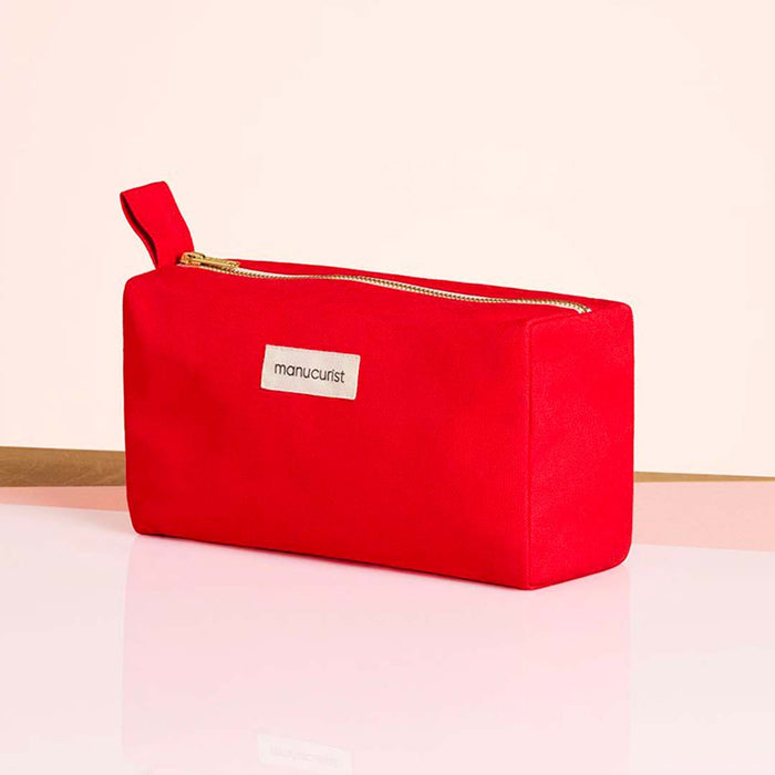The Manucurist-us Red Travel Bag, a rectangular organic cotton pouch with a zipper and side handle, stands on a glossy surface. A white label with the brand name is visible. The background is light pink with a wooden accent.
