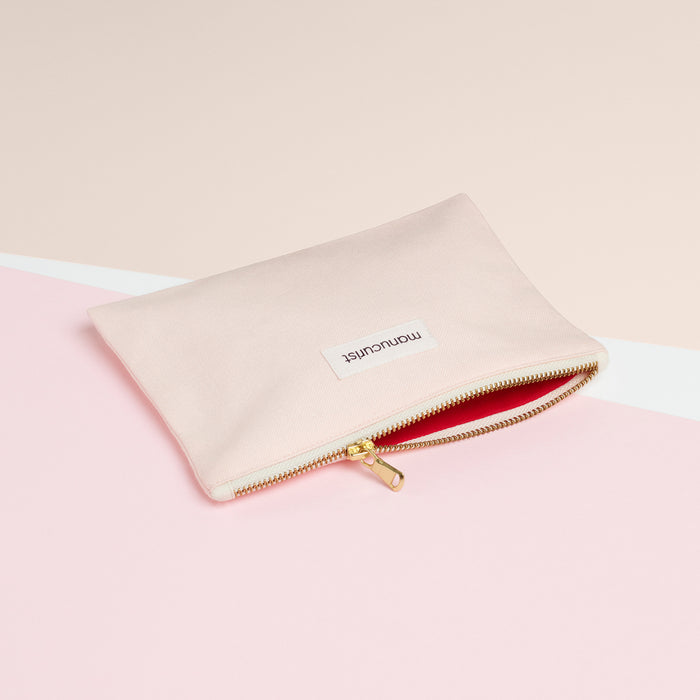 The Manucurist-us Small Pink Pouch, made from organic cotton with a gold zipper and a white “momentist” label, sits on a pink and cream background. Its red-lined interior is perfect for storing your beauty essentials.