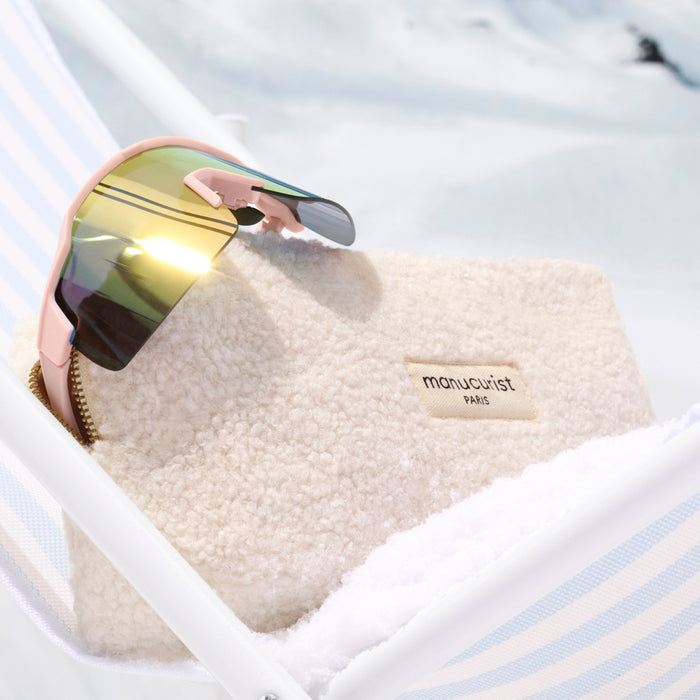 A pair of reflective sunglasses sits on the soft Teddy Bag by Manucurist-us, placed on a white and blue striped surface with a snowy backdrop—a chic winter accessory.