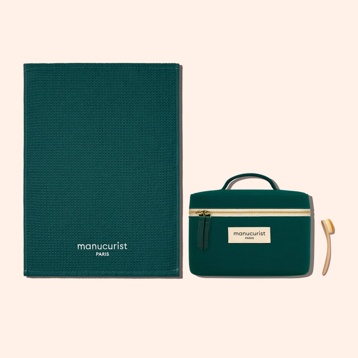 A dark green textured organic cotton Towel by Manucurist-us, a matching zippered pouch with handle and label, and a small wooden cuticle stick, all arranged on a light beige background for gentle surface protection.