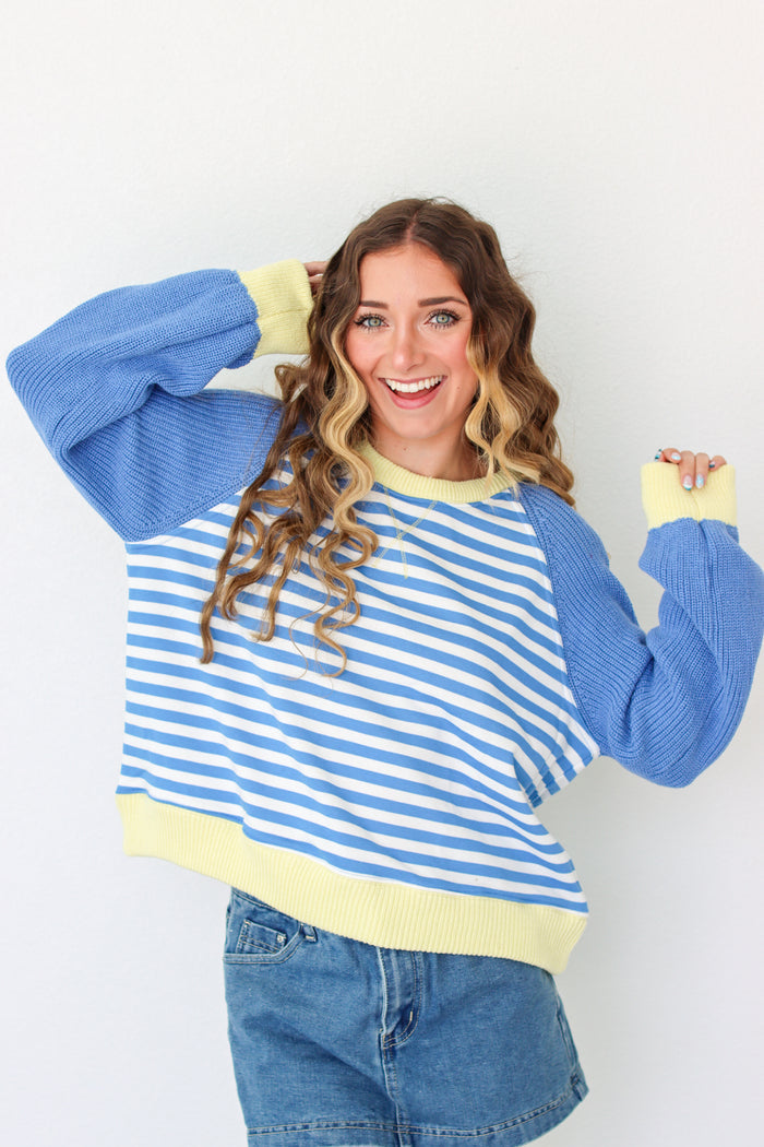 A woman modeling a blue and white striped knit sweater with solid blue sleeves and yellow ribbed cuffs and hem, paired with blue denim shorts against a plain white background.