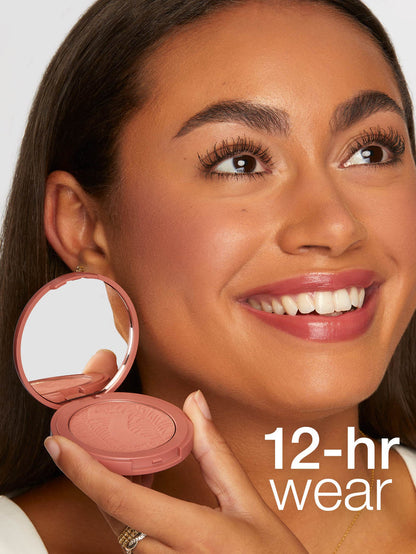 A smiling person holds an open compact blusher in hand, displaying its mirror and product. The text reads 12-hr wear on the right. The background is plain white.