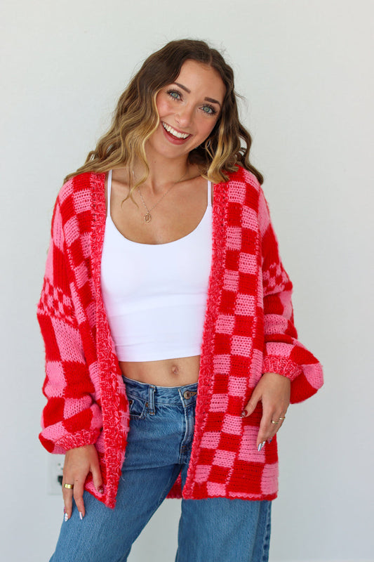 Woman wearing a red and pink checkered cardigan over a white top and blue jeans on a plain background
