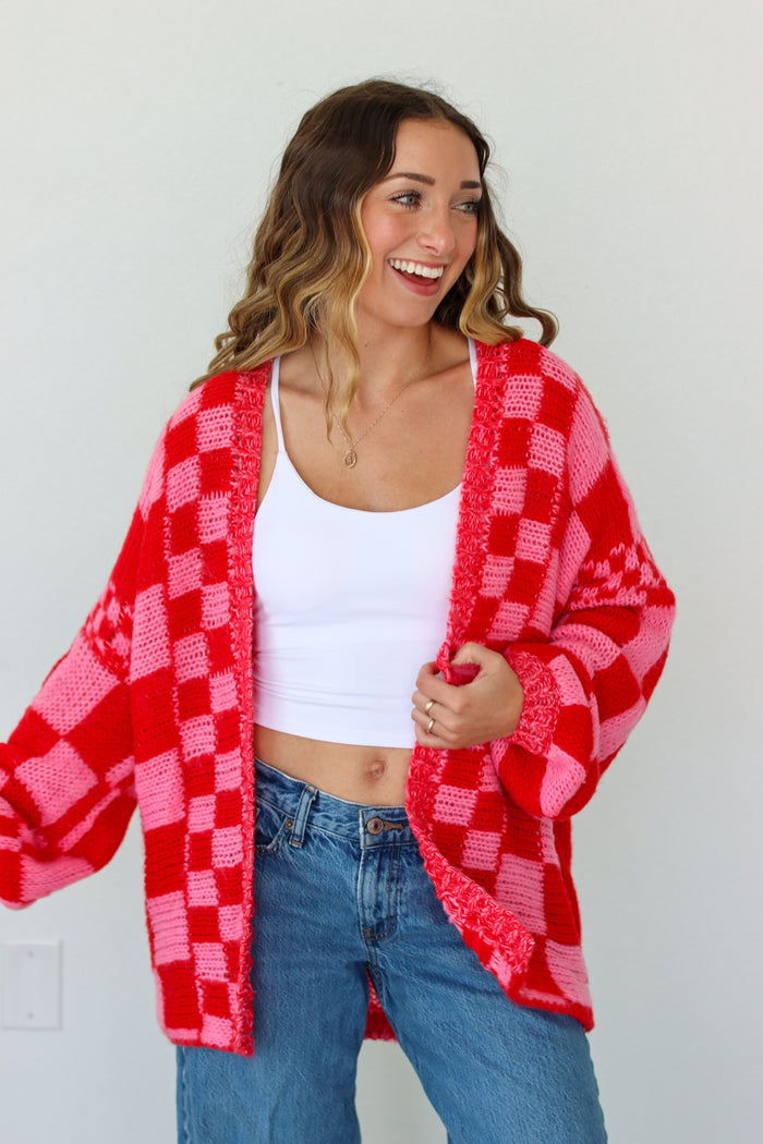 Woman wearing a red and pink checkered cardigan over a white top and blue jeans on a plain background