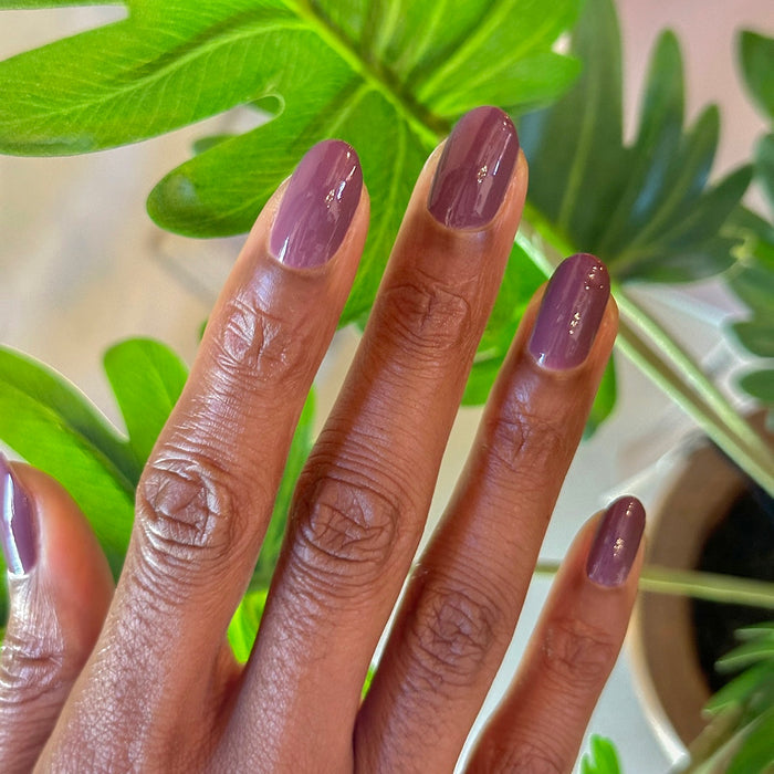 Hand with shiny gel polish manicure in Cardigan’s cozy true mauve shade against green leaf background