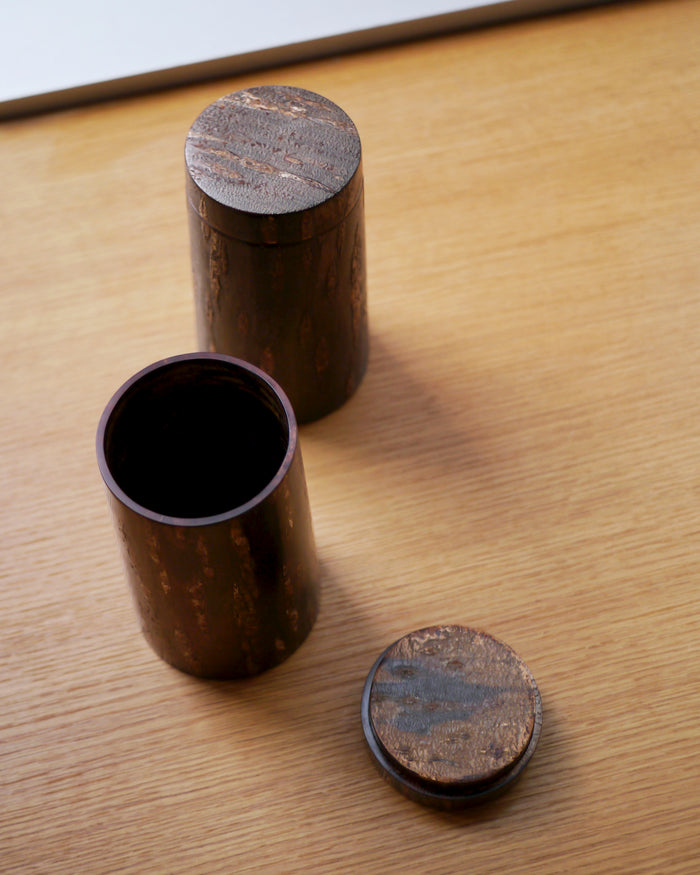 Two wooden cylindrical coffee containers with lids by Denshiro on a wooden surface