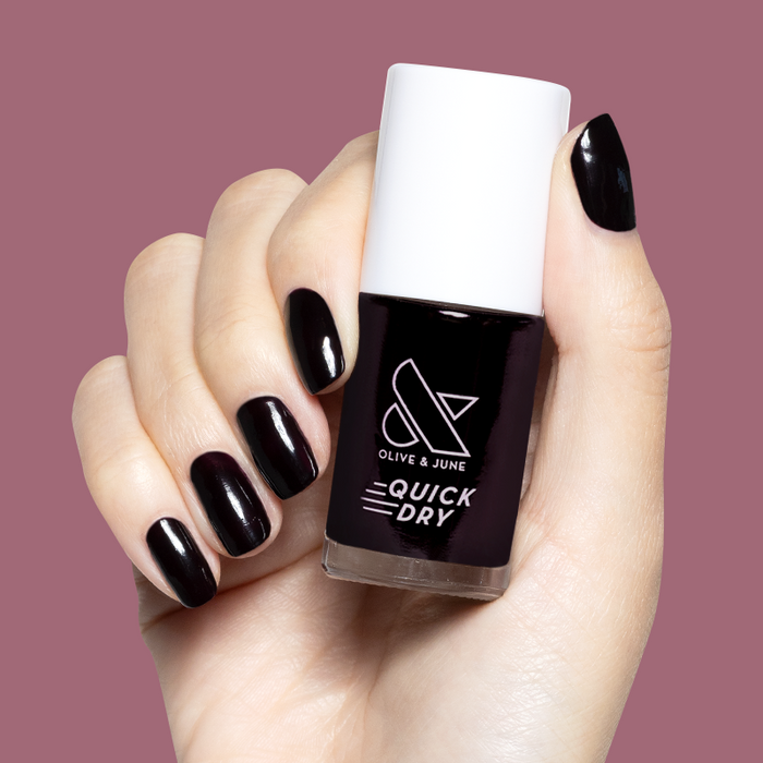 Hand holding Olive and June Covet quick-dry gel polish bottle with matching dark plum manicure on short nails