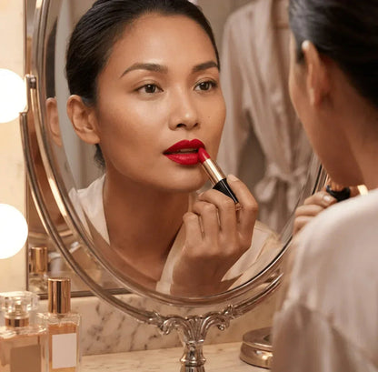 Woman applying bold red lipstick in front of a mirror on a vanity with perfume bottles.