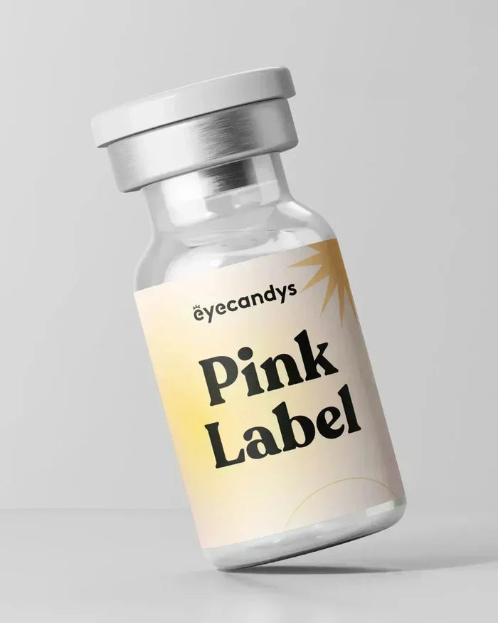 A small clear vial with a silver cap labeled "EyeCandys" is shown on a pastel yellow and white background. Inside are EyeCandys Attitude Glitter Pink color contact lenses, designed for comfort with high water content. The bottle rests against a gray backdrop.
