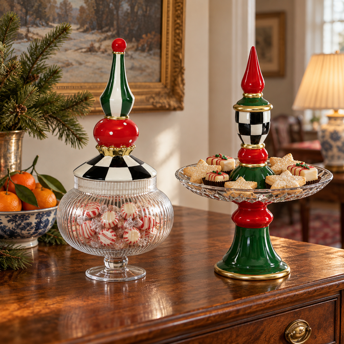 Two festive holiday-themed candy and snack stands on a polished wooden table in a warmly lit living room. One stand holds a glass jar filled with red-and-white striped peppermint candies, while the other is a tiered stand holding assorted pastries.