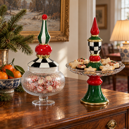 Two festive holiday-themed candy and snack stands on a polished wooden table in a warmly lit living room. One stand holds a glass jar filled with red-and-white striped peppermint candies, while the other is a tiered stand holding assorted pastries.