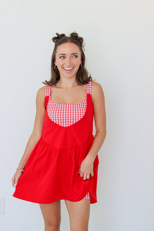 A woman modeling a red pinafore-style dress with red and white gingham trim on the bodice and straps, standing against a plain white wall.