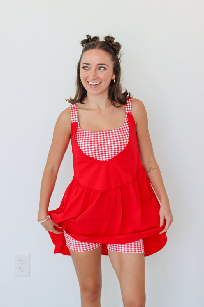 A woman modeling a red pinafore-style dress with red and white gingham trim on the bodice and straps, standing against a plain white wall.