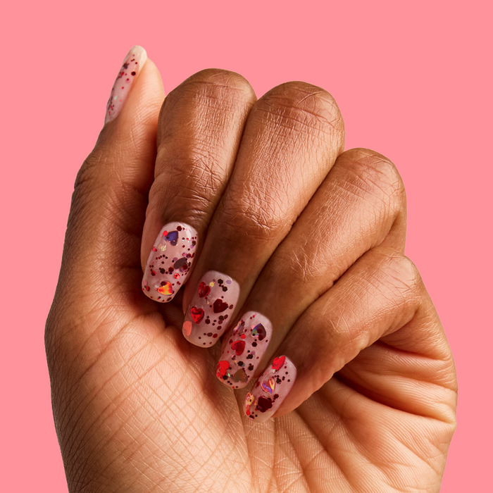 Hand showing nails with HEARTED quick dry polish featuring colorful heart and sequin confetti on a pink background