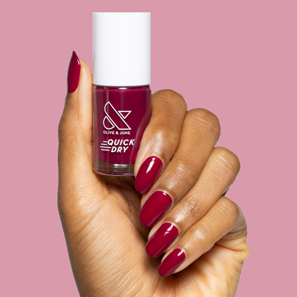 Hand holding Mulberry deep plummy quick dry gel polish bottle with matching manicure on natural nails against pink background