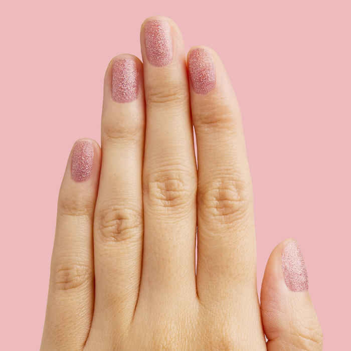Hand with nails painted in glittery pink holographic MWAH quick dry polish by Olive and June on pink background