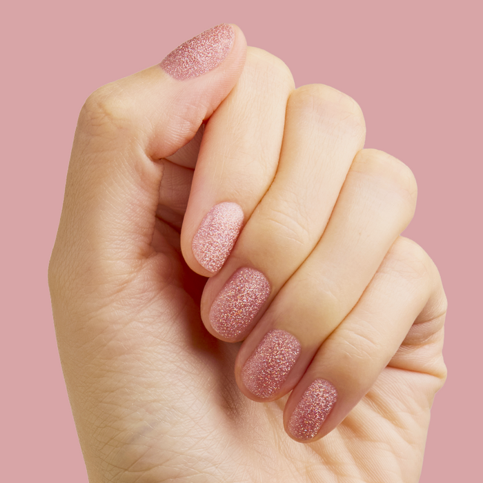 Hand with nails coated in pink holographic glitter MWAH quick dry polish by Olive and June on soft pink background