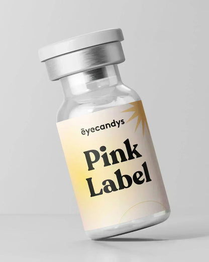 A small glass vial with a white cap labeled “EyeCandys Pink Label Angel Green” in bold black text rests on a pastel gradient background, perfect for storing natural color contact lenses. The backdrop is plain light gray.