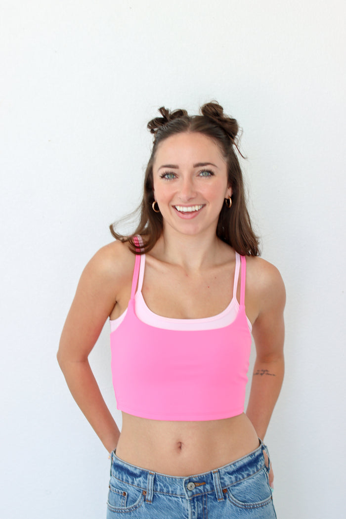 A smiling woman with her hair in two buns wears a bright pink crop top with a light pink trim, paired with blue jeans, standing against a plain white wall.