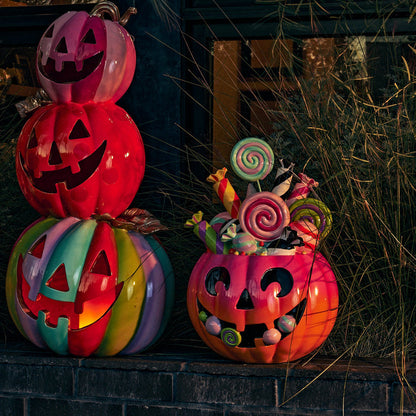 Three decorative jack-o'-lantern pumpkins with candy-filled tops are arranged on a dark brick ledge outdoors at dusk, with tall grasses and a building window in the background.