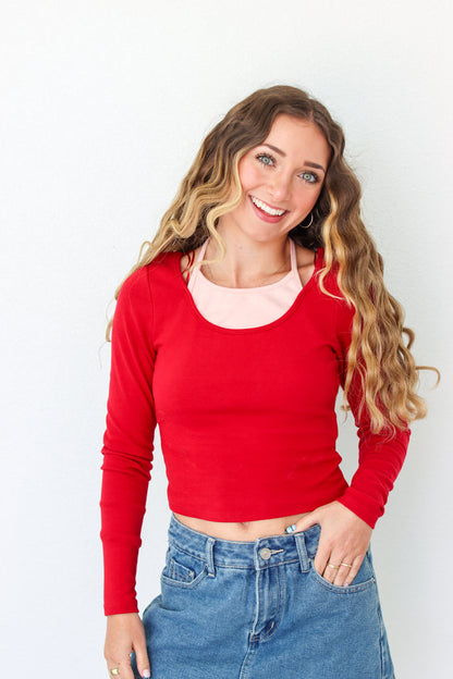A smiling woman with long wavy blonde hair wearing a bright red long-sleeve scoop-neck crop top over a light pink undershirt, paired with blue jeans, against a plain white background.