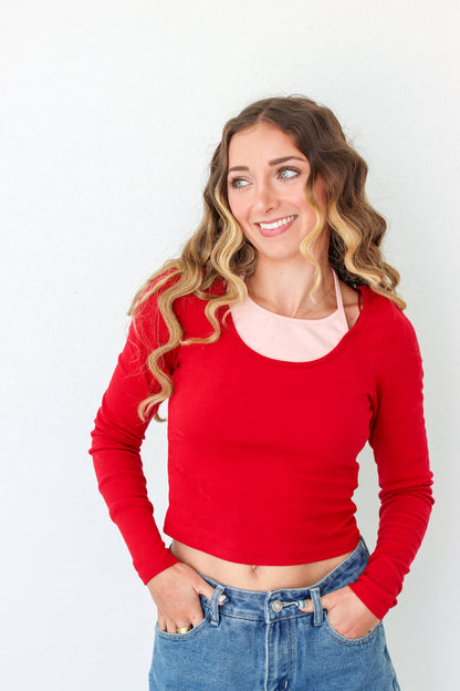 A woman with wavy blonde hair wearing a bright red long-sleeve crop top over a light pink undershirt and blue jeans, standing against a plain white background.