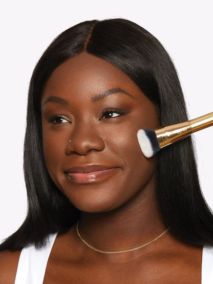 A woman gently applies makeup using a Tarte Cosmetics brush, smiling slightly. She wears a white top and a thin necklace, set against a plain white background.