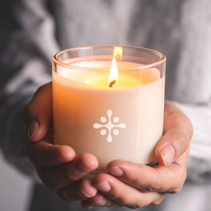 SKN072 SKNLOGIC Pineapple Soy Body Massage Candle | A person's hands holding a lit candle in a clear glass jar, with a white geometric flower-like logo printed on the front. The background is a soft, out-of-focus gray fabric.