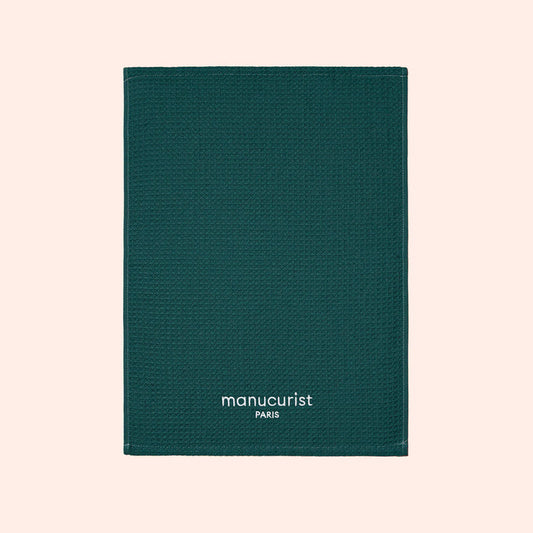 A green textured towel from Manucurist-us, featuring white "manucurist PARIS" text at the bottom center, displayed on a light beige background and ideal for surface protection.
