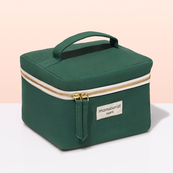 A green fabric Manucurist-test Vanity Case with a handle, gold zippers, and a beige "manucurist PARIS" label—ideal for nail polish or manicure storage—shown on a white surface with a light pink background.