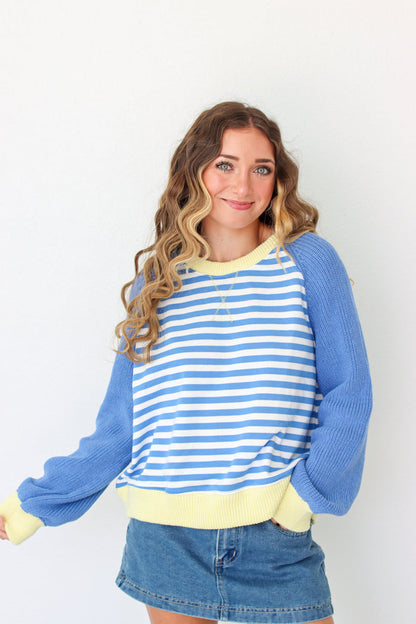 A woman wearing a blue and white striped long-sleeve sweater with solid blue sleeves and a pale yellow ribbed collar, hem, and cuffs, paired with a denim skirt against a plain white background.