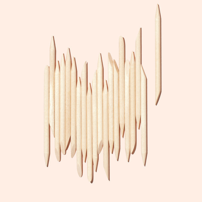 A set of 20 Manucurist wooden cuticle sticks is arranged vertically on a light pink background, with one stick separated to the right, showcasing their nail cleaning use.