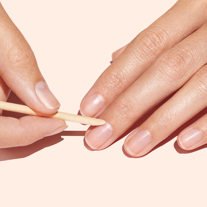 A hand uses a Manucurist Wooden cuticle stick x20 to gently push back another hand’s cuticles against a light peach background, as part of a nail cleaning routine.